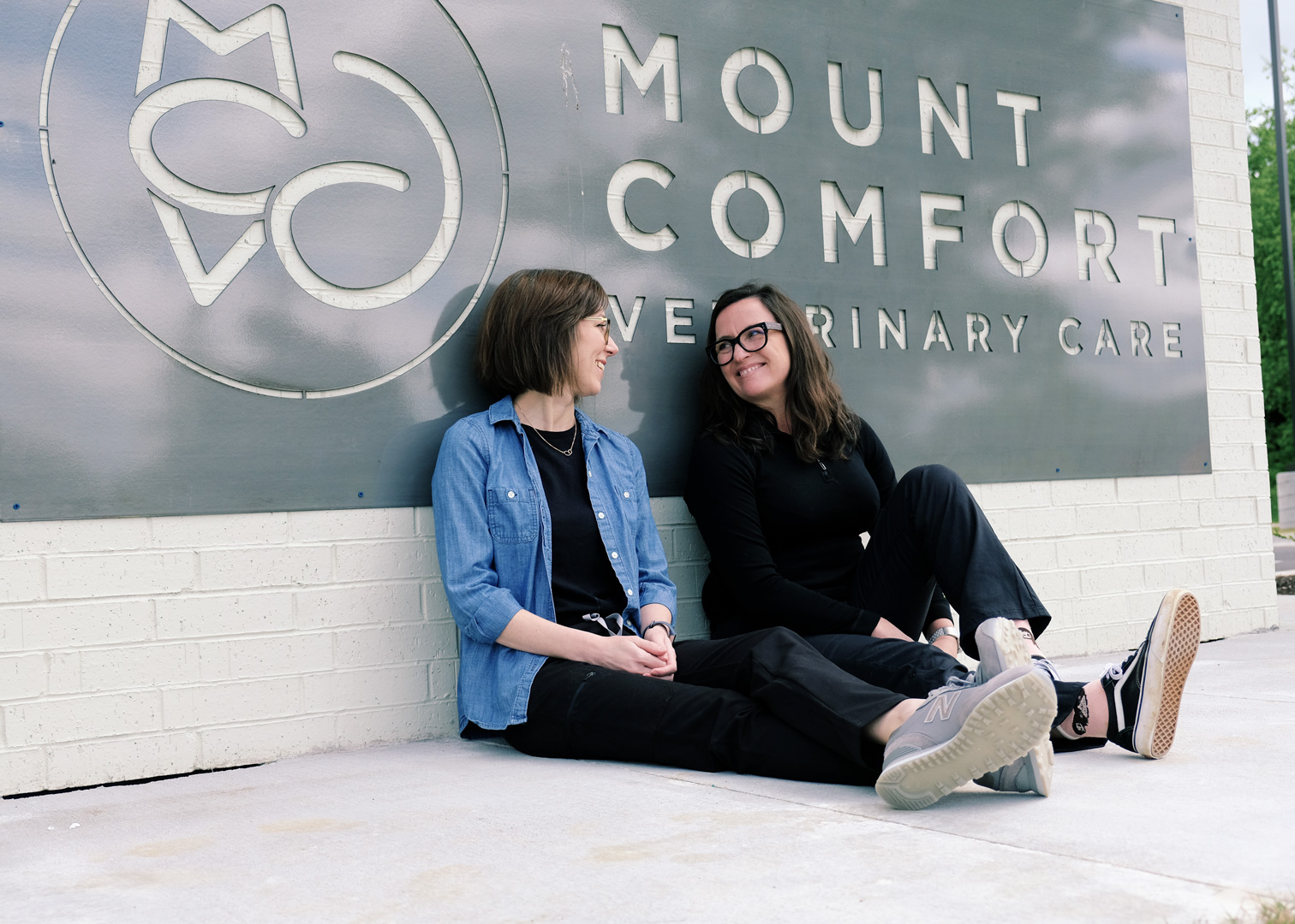 Dr. Heather Williams and Dr. Sarah Hantz sitting in front on the cut metal Mount Comfort Veterinary Care sign on the clinic wall